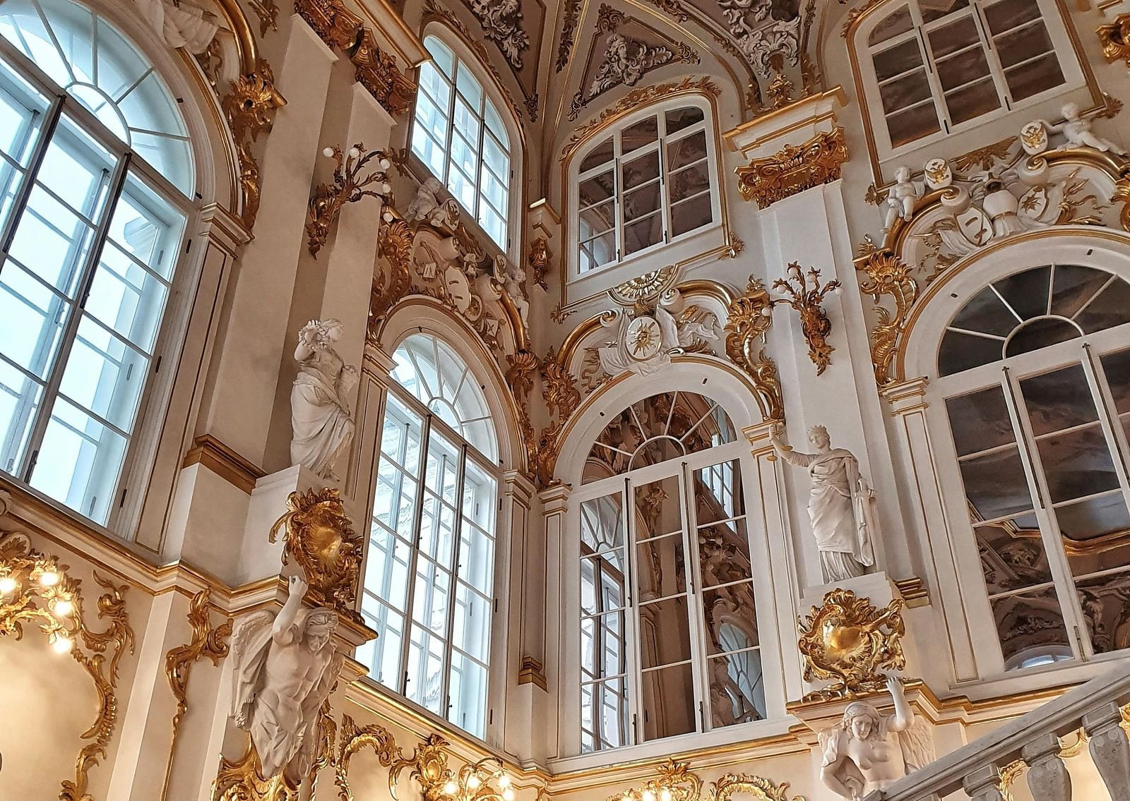 A low-angle shot of a magnificent staircase in a palace, with gold and white walls adorned with intricate carvings, large windows, and a beautifully painted ceiling.