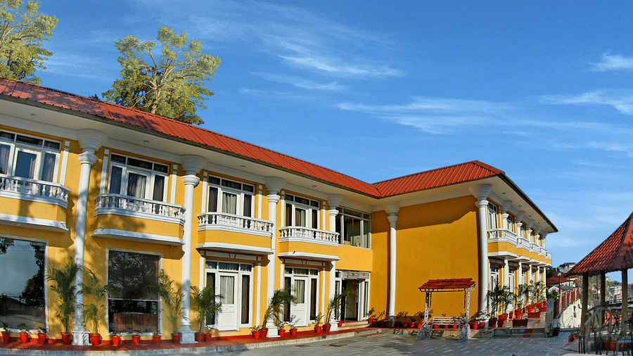 A corner exterior view of the  building with a roof and a balcony, surrounded by trees and a clear blue sky.
