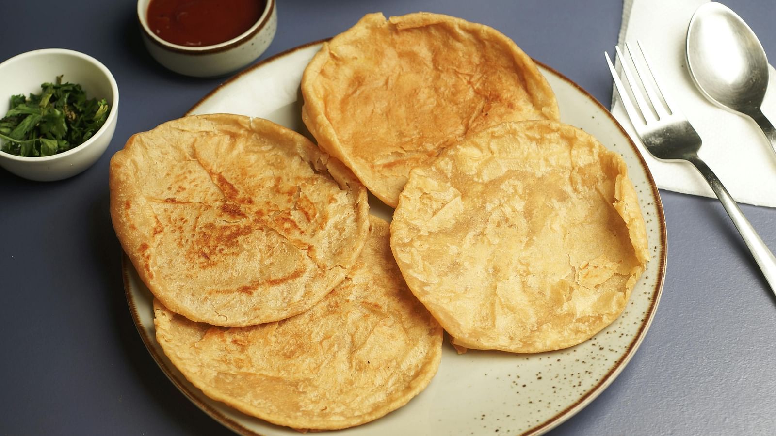 A traditional Indian meal consisting of golden flatbreads served with side dishes including chutney, a vegetable curry, and pickle on a grey table.