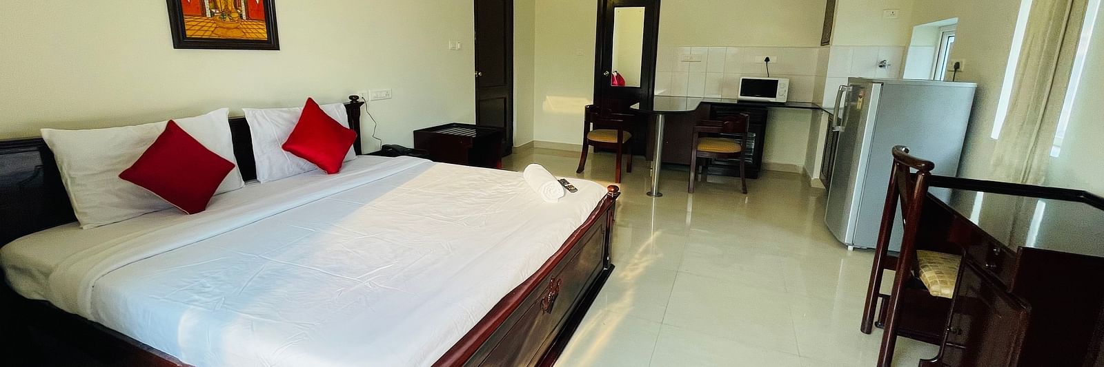 A room with a double bed featuring white linens and red cushions, a separate seating area, and a glimpse into a kitchenette space at Sanctum Suites Indiranagar, Bangalore.