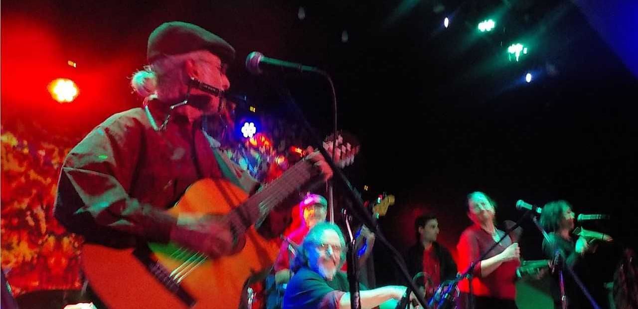 A musician from the Pura Vida Band performing on stage, playing a guitar under colorful lights, with the band members in the background.