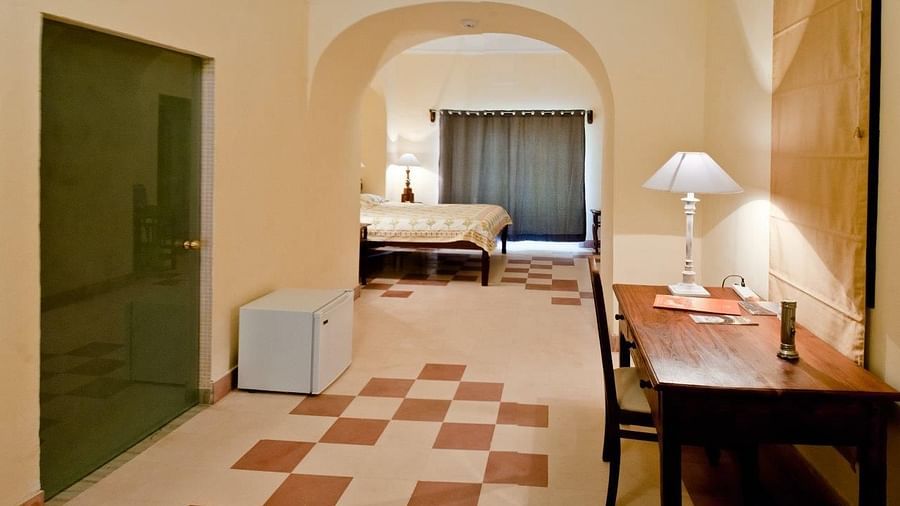 Hotel hallway with a desk and chair, a mini-fridge, a doorway, and an arched opening leading to a bedroom with a bed at Tijara Fort-Palace, Alwar.