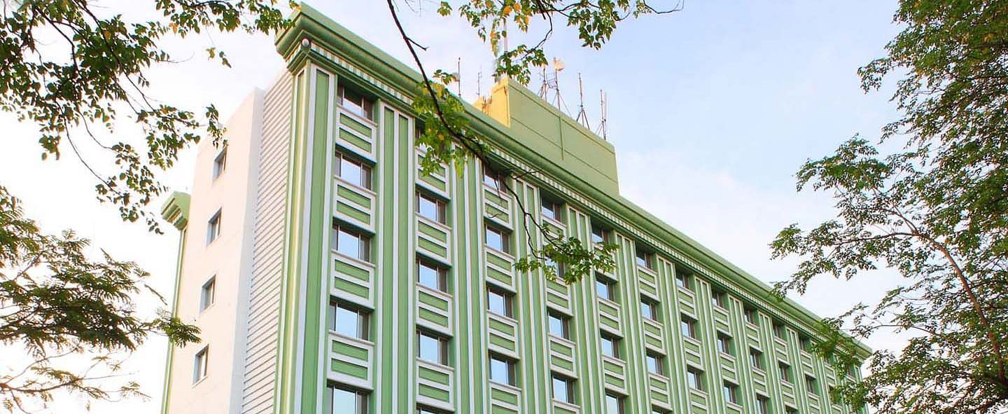 A multi-story green hotel building flanked by trees at Tara, Comfort Hotel in Hyderabad - Ramoji Film City.