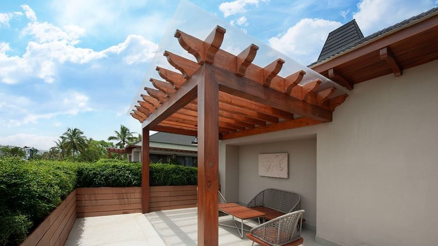 A modern and relaxing private balcony at The Evren, Vagator. The space features two comfortable chairs and a table under a stylish wooden pergola with a clear roof, offering a view of the surrounding greenery and a clear blue sky.