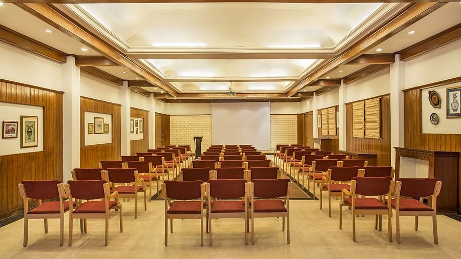 A spacious conference room with rows of chairs facing a projection screen. The room has high ceilings, wooden paneling, and framed artwork on the walls. The overall ambiance is professional and formal - the naini retreat, nainital.