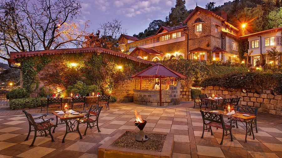 A beautiful outdoor dining area at the naini retreat, nainital. There are tables and chairs set up on a stone patio, with a fireplace in the center. The building is lit up at dusk, creating a warm atmosphere. 