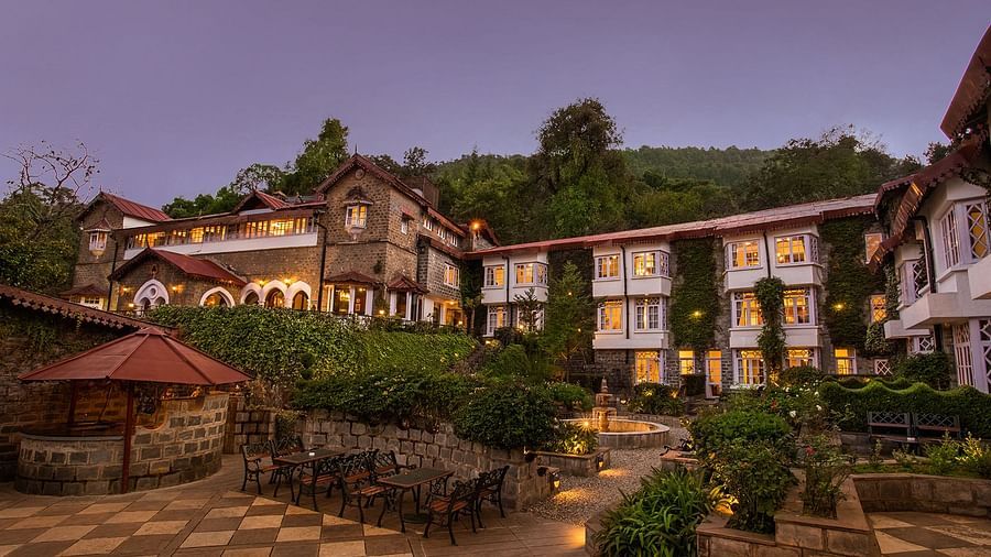 An image of the naini retreat, nainital. features multiple buildings with chairs in exterior with tables and gardens creating a serene atmosphere, surrounded by hills, captured in the evening.