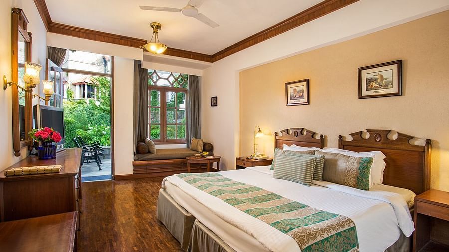 A well decorated room with a large bed, a shiny mirror alongside a make up table, with multiple wall decors and lights, portrayed while showcasing the lush outdoors through the doorhole- The Naini Retreat, Nainital.