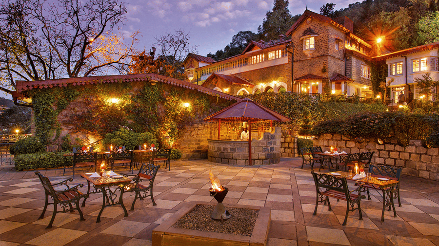 A beautiful outdoor dining area at the naini retreat, nainital. There are tables and chairs set up on a stone patio, with a fireplace in the center. The building is lit up at dusk, creating a warm atmosphere. 