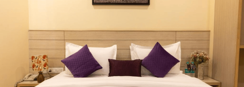 A hotel room, featuring a bed with white linens and a purple runner, a small framed painting above the bed, and a small statue on the nightstand | Perfectstayz Value Sagar