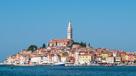 Coastal town with terracotta-roofed buildings and a tall bell tower, set against a deep blue sea and clear sky.