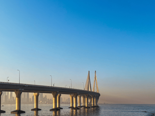 A long sea bridge stretches across calm waters, framed by a clear blue sky and rocky shoreline in the foreground.