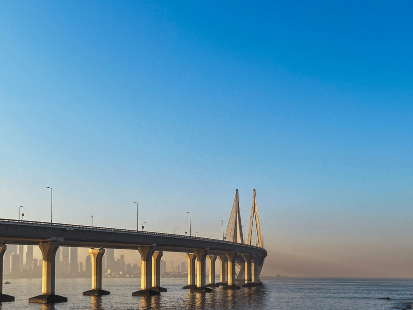 A long sea bridge stretches across calm waters, framed by a clear blue sky and rocky shoreline in the foreground.