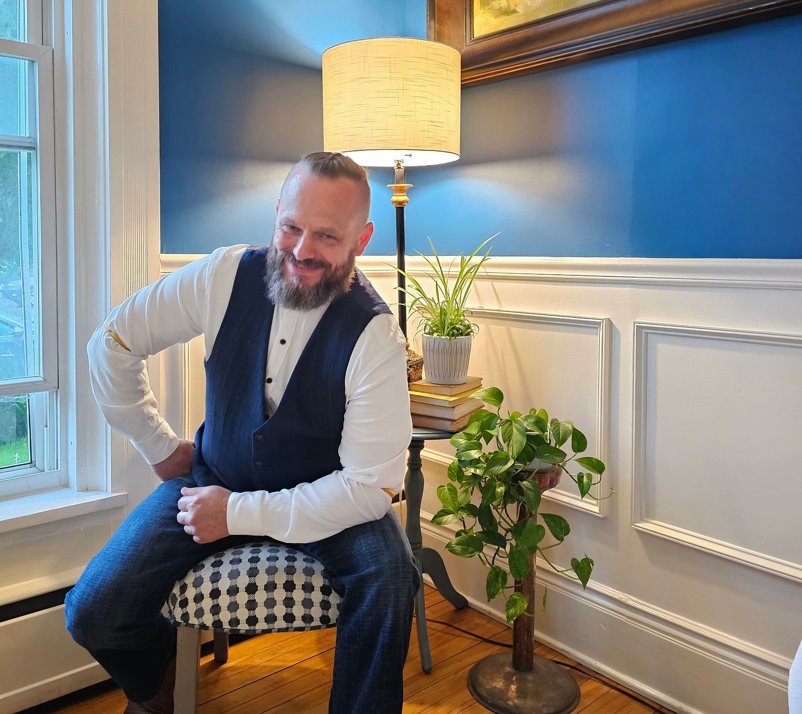 Chef Tom E. White seated by a window with natural light, holding papers, dressed in a vest and white shirt.