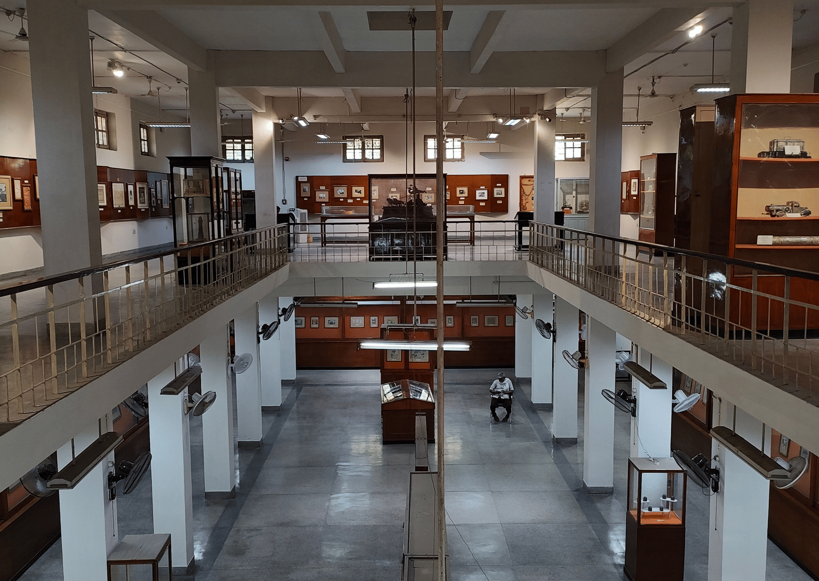 Interior of a museum with white columns, gray floors, display cases, framed exhibits along the walls, and a mezzanine level surrounding an open central space.