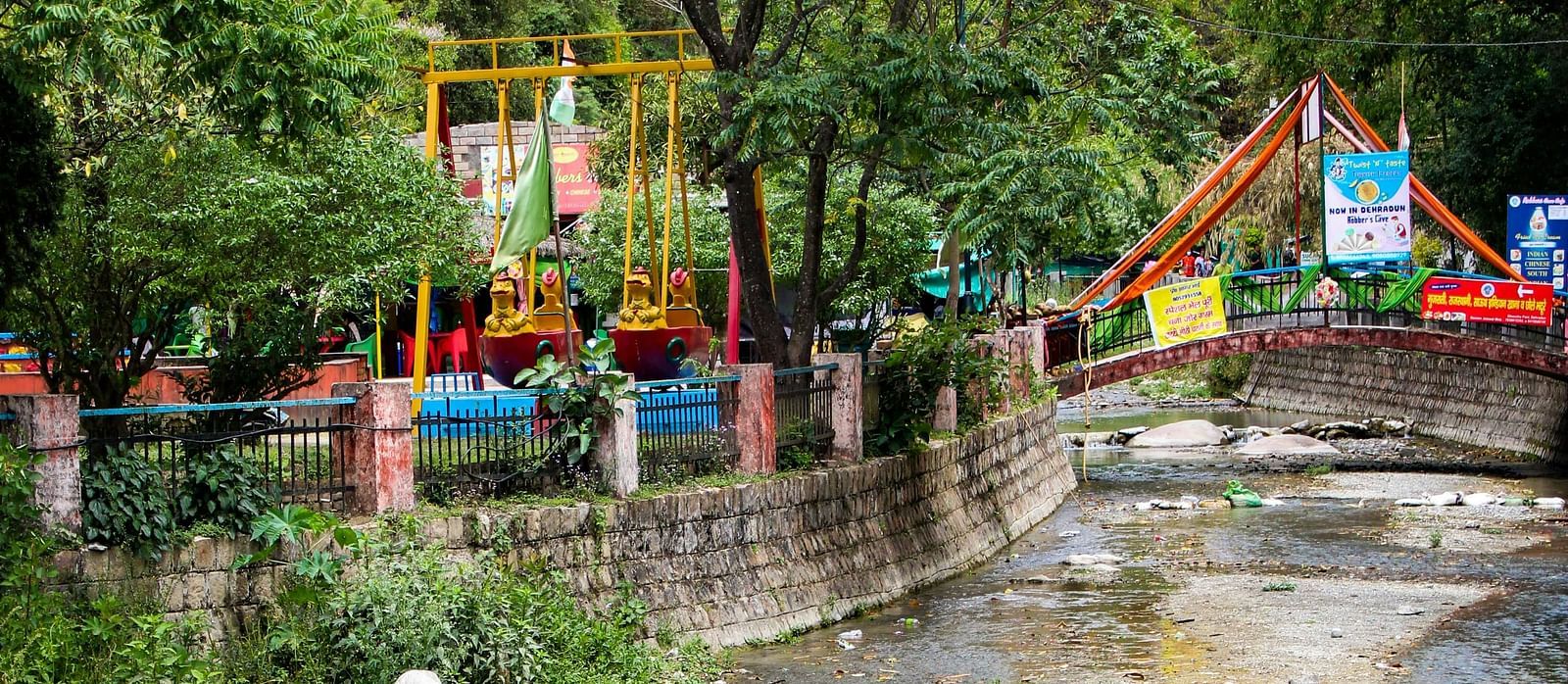 A small, playful waterfall flowing under a pedestrian bridge in a park, with surrounding greenery and recreational structures.