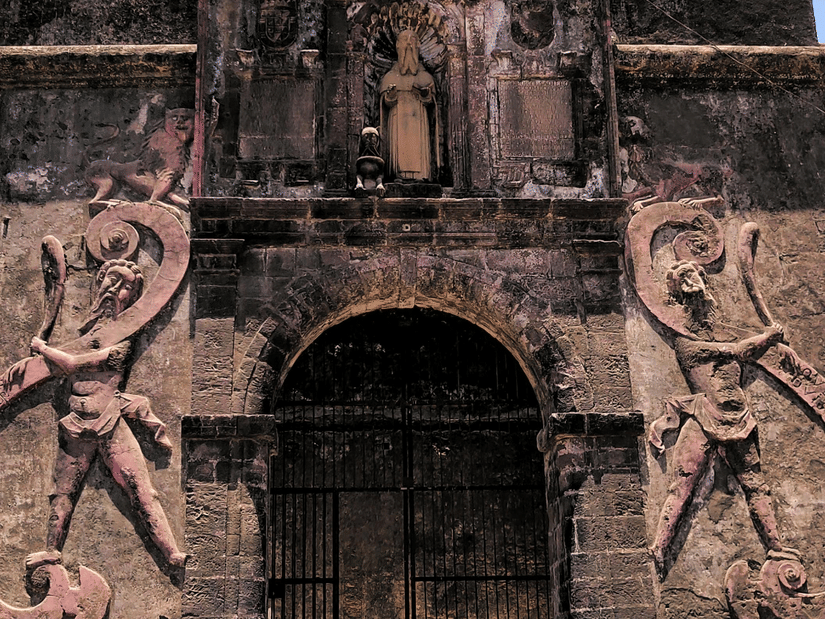 The entrance of an old stone fort, featuring intricate carvings and a cross at the top of the structure, set against a clear blue sky.