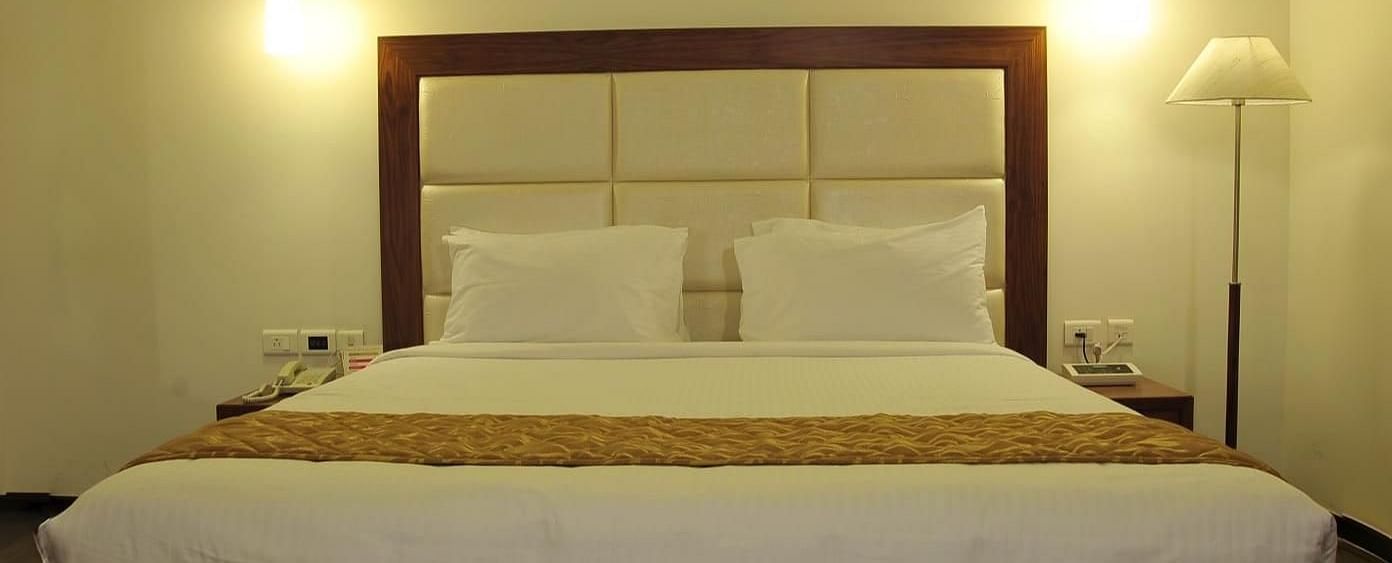 A queen size bed with white linens and a gold runner, two hanging lights, a floor lamp, and a white rug on a wooden floor at Dhanalakshmi Srinivasan Hotels, Perambalur.