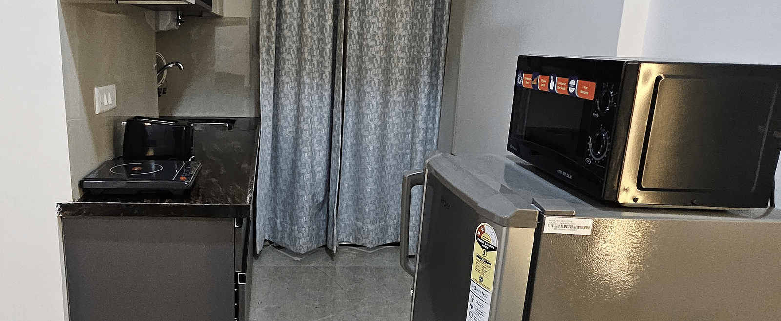 Compact kitchenette in a studio room featuring a refrigerator, microwave, induction cooktop, sink, and overhead cabinets with a curtain partition - Urban Suites by BluSalzz, Jaipur