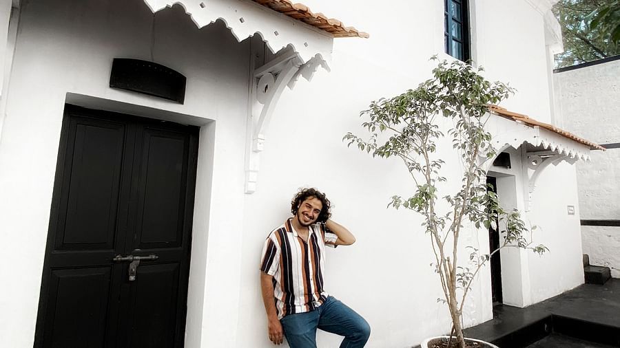 A person in a striped shirt and light-coloured pants leans against a white building with dark doors and windows, at Fort Tiracol, Goa. 