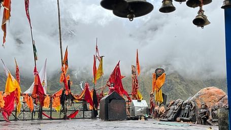 Misty Himalayan temple courtyard with colourful prayer flags, hanging brass bells, and wet stone steps leading up to carved idols and cloud-covered hills.