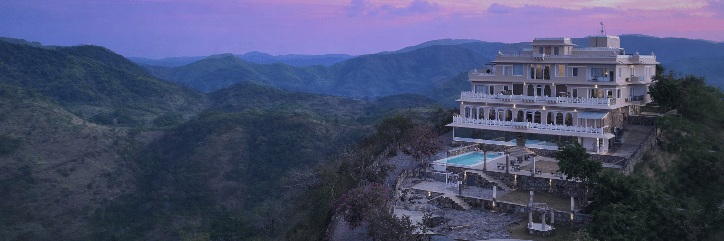 Facade of Fateh Safari Suites, Kumbhalgarh featuring multiple storeys, swimming pool, lawn, sitting area, and more amidst the green hills.
