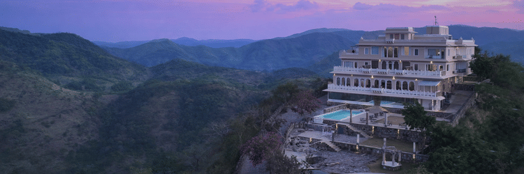 Facade of Fateh Safari Suites, Kumbhalgarh featuring multiple storeys, swimming pool, lawn, sitting area, and more amidst the green hills.