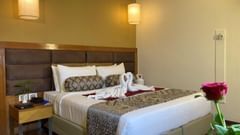 A hotel room with a bed and decorative towels, a nightstand with a lamp, and a red rose on a small table.