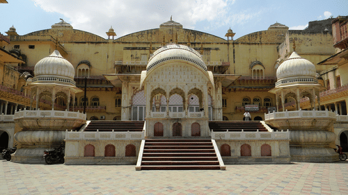 The Alwar City Palace featuring a grand facade with domes, arched doorways, and a wide paved courtyard under a partly cloudy sky.