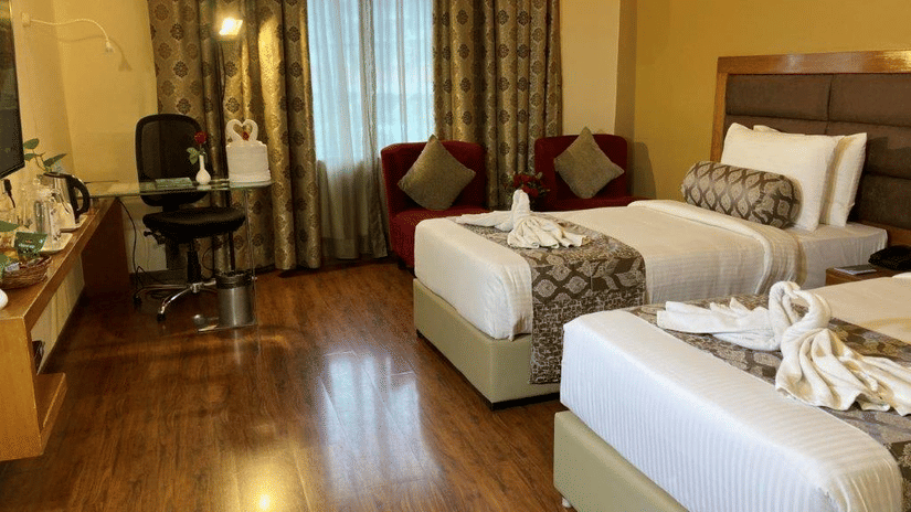 A room with twin beds, wooden flooring, two sofas and a work desk with a window behind.