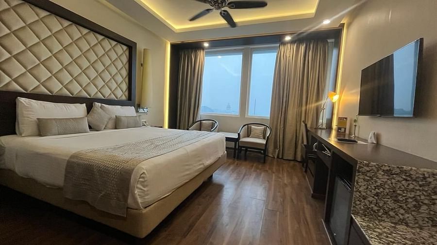 Image of a hotel room in ambient warm lighting featuring plush bed with a TV counter in front of it and a huge window next to the sitting space