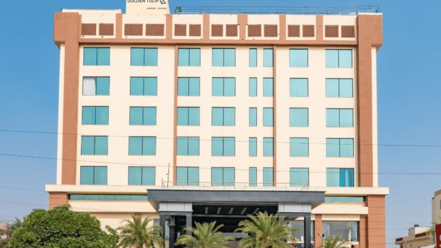 A modern, multi-storey hotel with a beige facade and palm trees at the entrance. Bright, clear daylight with a blue sky background at Golden Tulip Kukas, Jaipur.