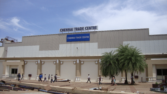 Facade of Chennai Trade Fair with two trees in front of it and people walking in front of it.