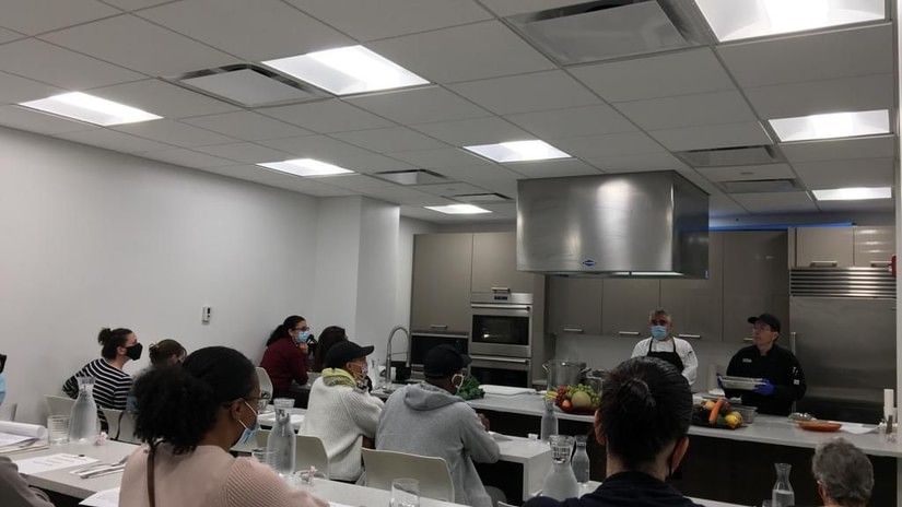 A group of people attending a cooking class, watching chefs demonstrate techniques at the front of a modern kitchen classroom at YO1 Longevity & Health Resorts, Catskills