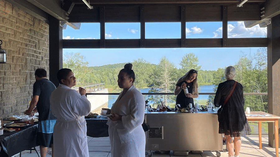 People standing on a patio overlooking a lake on a sunny day, and some serving themselves food from the counters at YO1 Longevity & Health Resorts, Catskills