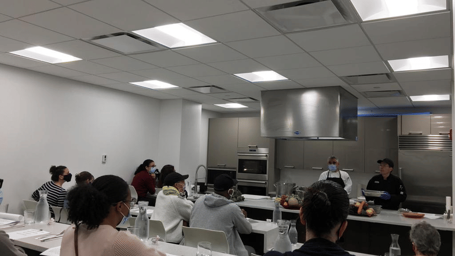 A group of people attending a cooking class, watching chefs demonstrate techniques at the front of a modern kitchen classroom at YO1 Longevity & Health Resorts, Catskills