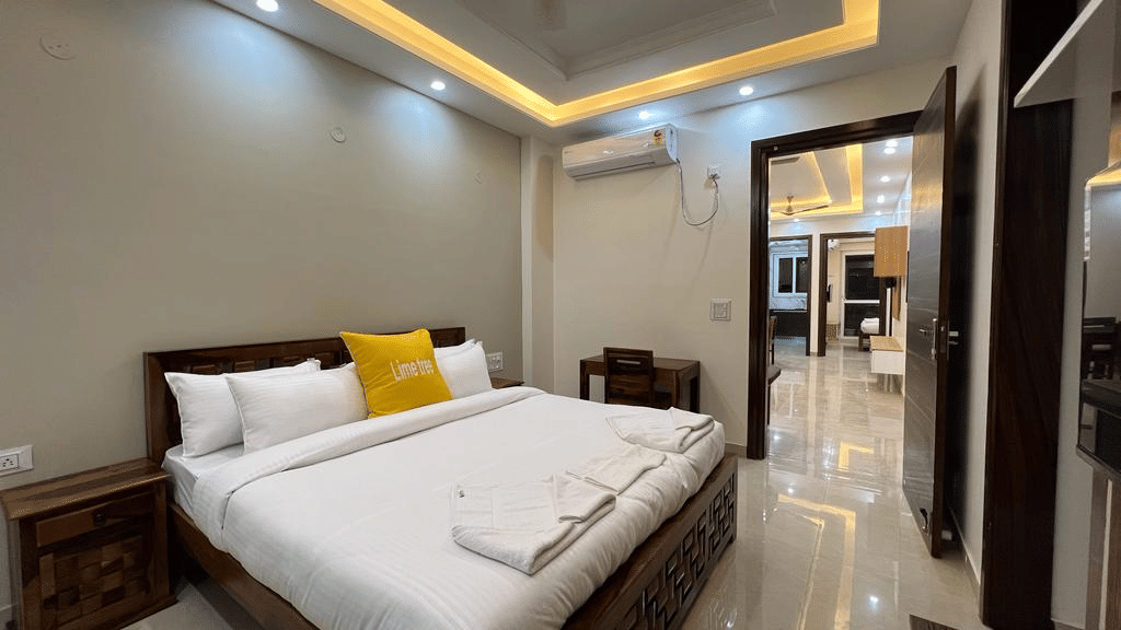 Bedroom of Lime Tree Hotels & Service Apartment Private Limited featuring a white bed with a yellow accent pillow and an open view towards an adjoining room.