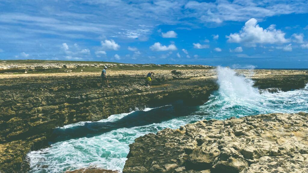 Ocean waves crash dramatically against rugged, dark rock formations under a partly cloudy blue sky, with distant figures on the landscape - Tamarind Hills Resort and Villas - Antigua