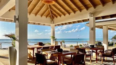 Open-air seaside restaurant with wooden furniture and a high thatched roof, overlooking a sunny ocean view under a clear blue sky.