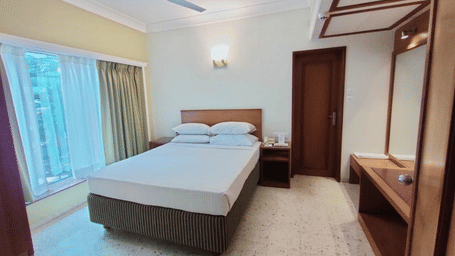 A bedroom containing a double bed with white linen, a ceiling fan, a large window, and wooden furniture | Trance Oakwood Hotel, Pune