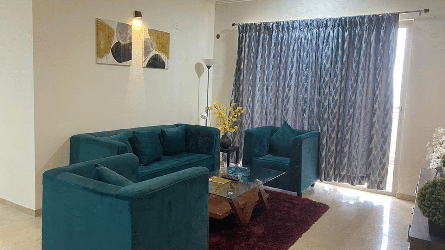 A stylish living area with a teal velvet sofa set, a glass coffee table over a maroon rug, and patterned curtains in a modern suite at Apartel by Aarin, Oragadam.