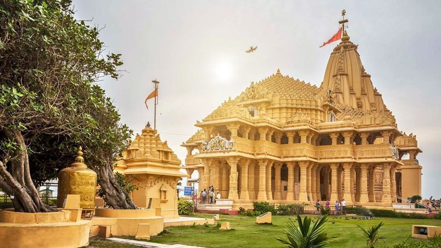 A grand sandstone Somnath temple with ornate carvings and fluttering saffron flags, surrounded by lush greenery.