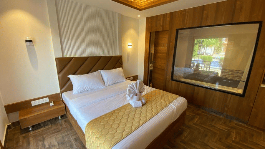 Premium resort bedroom with a quilted brown headboard, wooden paneling, and a large window looking into the bathroom - DAN Resorts & Weddings, Palghar