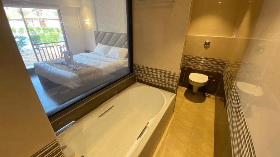 Luxury bathroom featuring a large white soaking tub and a glass partition looking into the main bedroom - DAN Resorts & Weddings, Palghar