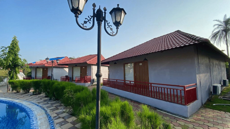 Rows of grey and red-roofed resort cottages situated along a curved blue swimming pool with vintage lamp posts - DAN Resorts & Weddings, Palghar