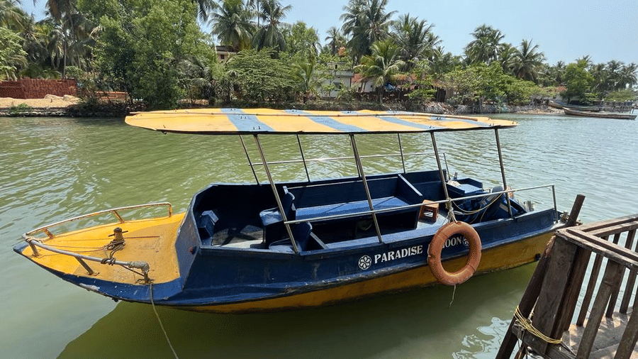 A yellow and blue motorboat docked beside a wooden jetty, surrounded by palm trees along the riverbank at Paradise Lagoon Resort, Udupi.