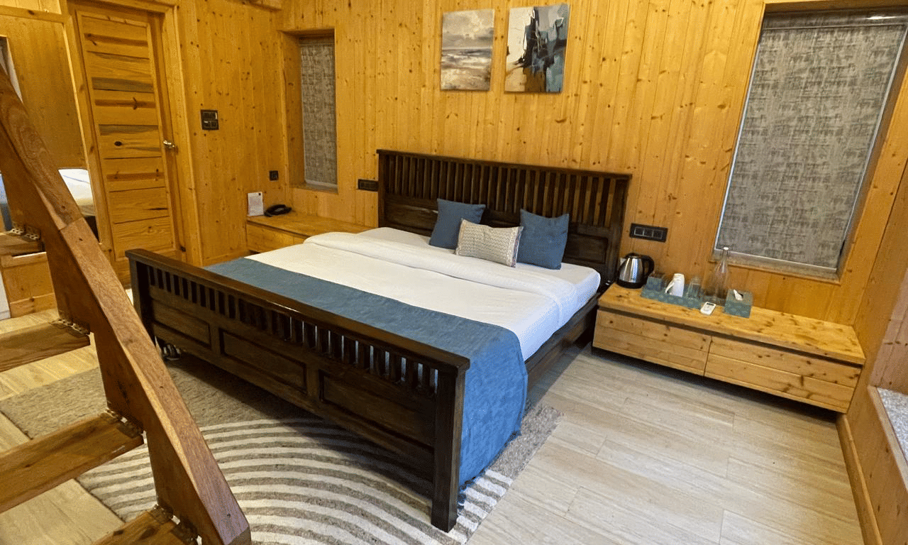 Megha Resort & Villas, Dehradun Standard Room, featuring a dark timber bed frame, timber-clad walls, a smooth light floor, and an internal wooden staircase leading to another level.