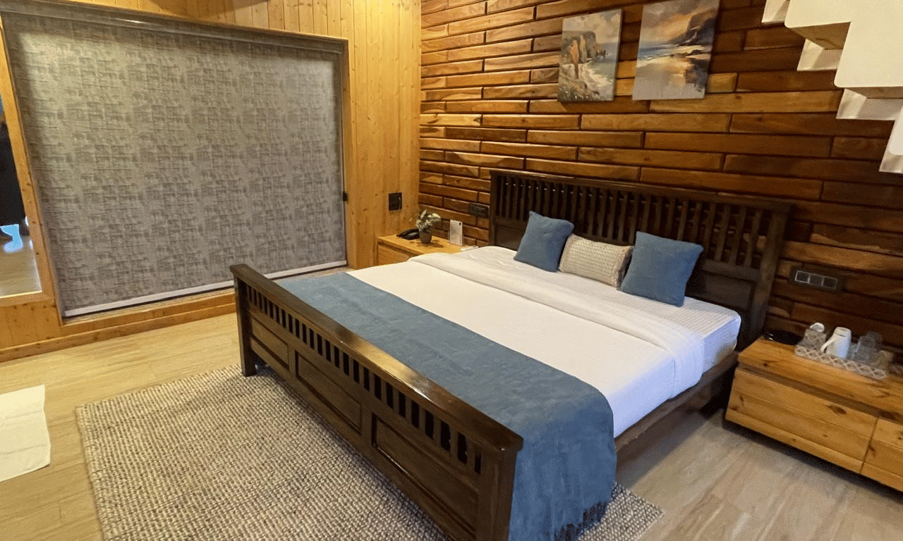 Megha Resort & Villas, Dehradun Standard Room, presenting a spacious interior with a prominent timber accent wall, a large bed with a fabric throw, and a textured area rug on a light wooden floor.