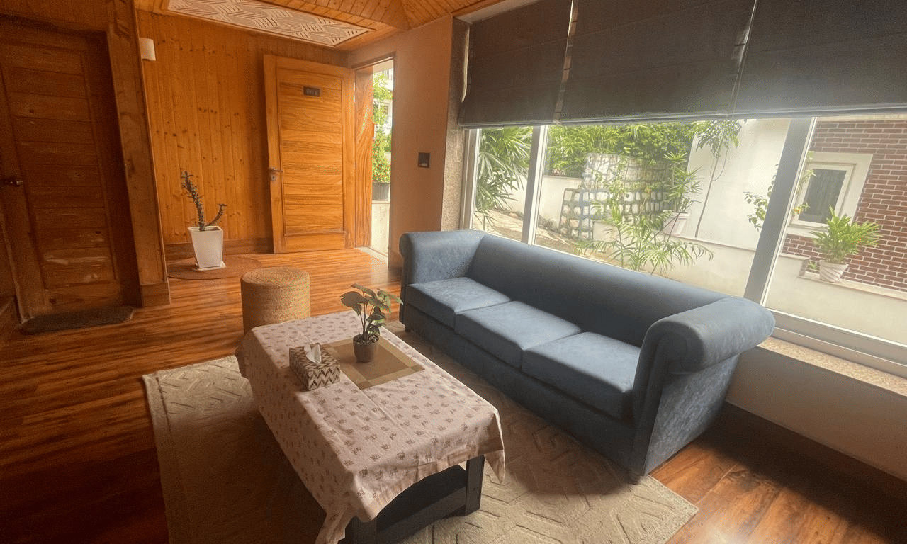 A cosy living room with wooden walls, a dark sofa, and large windows with blinds partially drawn at Megha Resort & Villas, Dehradun.