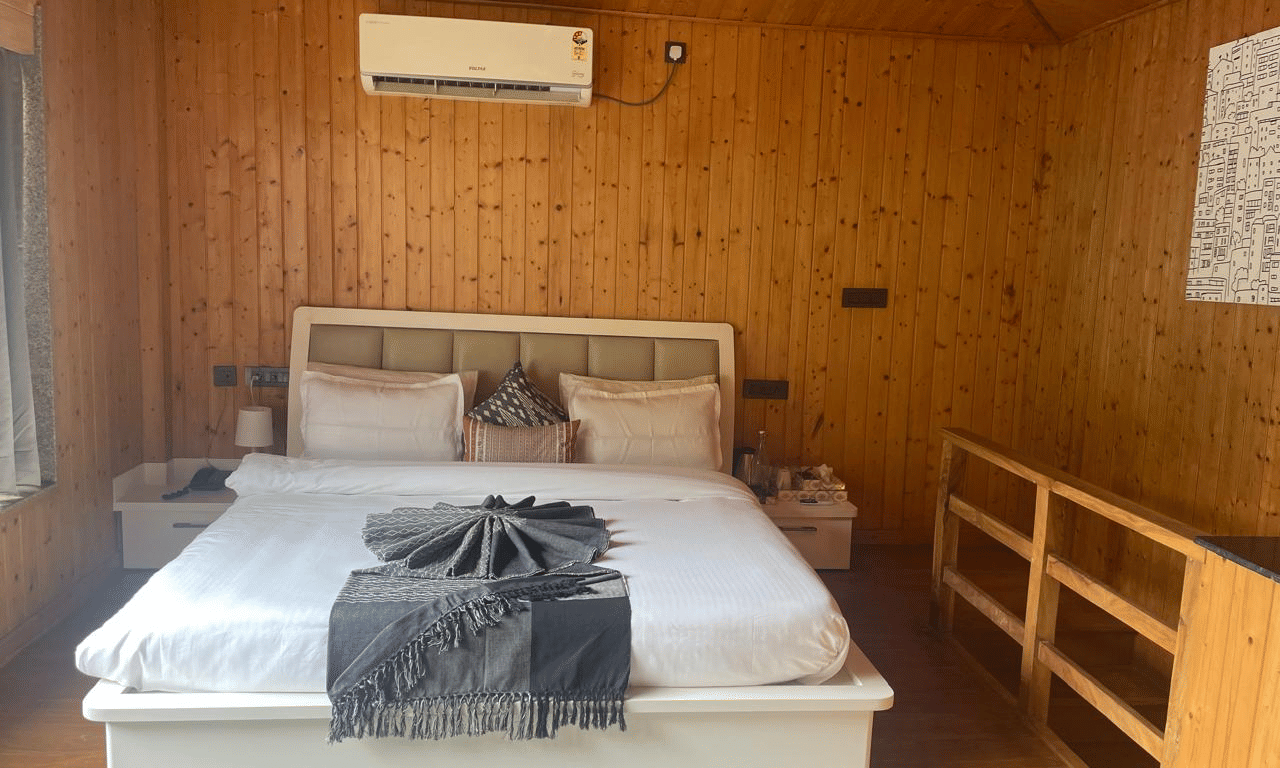 A wood-panelled bedroom with a white bed contrasts with a dark grey blanket and pillows at Megha Resort & Villas, Dehradun.